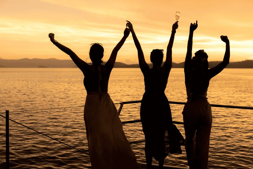 Three silhouetted figures with raised arms against a sunset over a body of water. 3