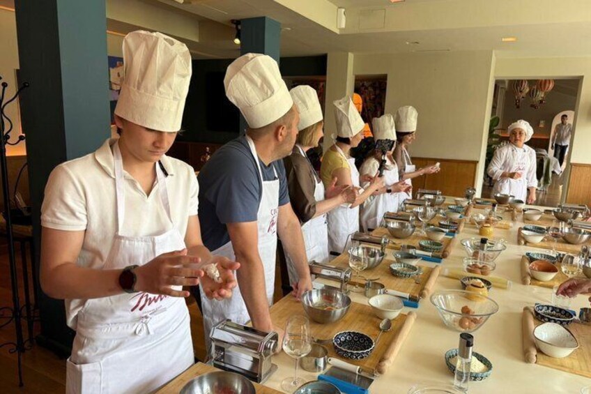 A group of chefs in aprons and hats preparing food at a long table with various cooking utensils and ingredients. 3