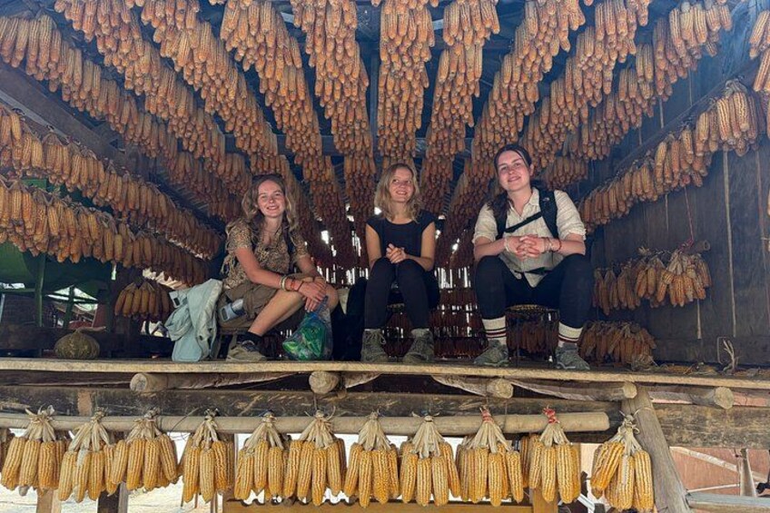 Three people sitting under a structure with hanging corn cobs. 3
