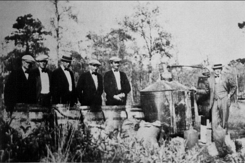 A group of men in formal attire standing outdoors, with a large metal container and a smaller structure in the background. 3