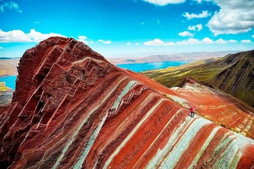 Una persona haciendo senderismo sobre una colorida formación rocosa a rayas, con un lago y montañas al fondo. 1