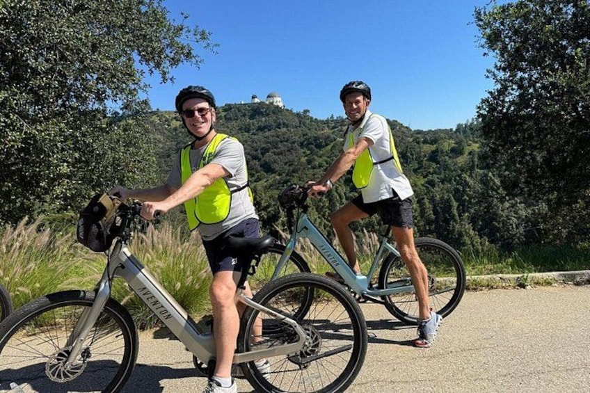 Two cyclists on a paved path with trees and hills in the background. 1