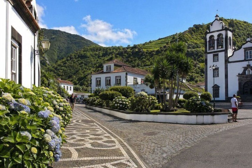 A cobblestone street lined with white buildings, a church with a bell tower, and lush greenery in the background. 3
