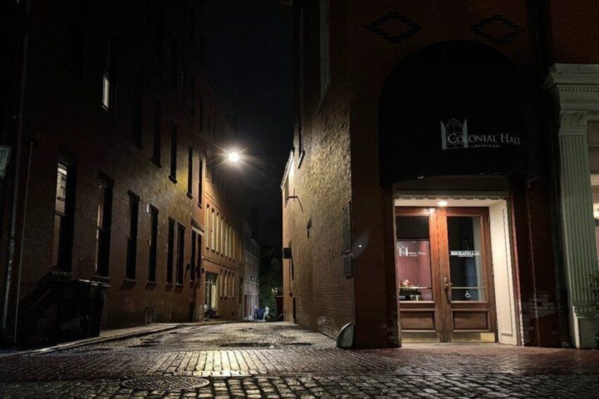 A nighttime street with cobblestones, a building with a sign reading 'Colonial Hall,' and a street lamp. 5