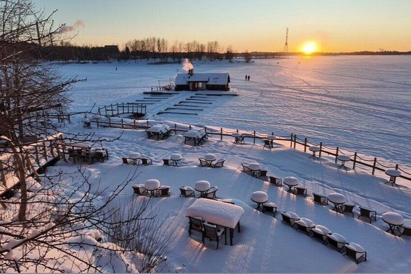A snow-covered outdoor area with tables and benches arranged in a semi-circle, a building in the background, and a sunset over a frozen body of water. 3