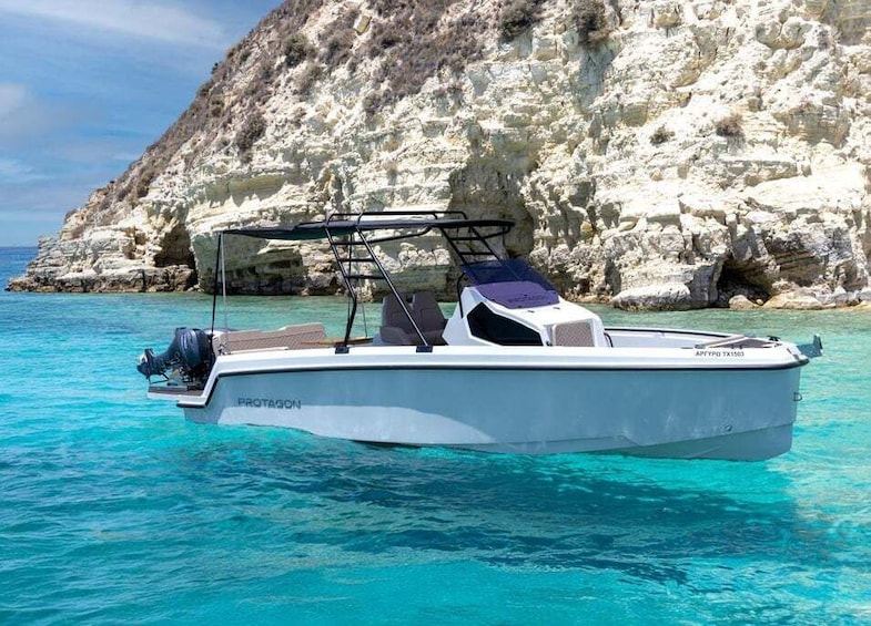 A white motorboat named 'PROTAGON' with a cabin and outboard motor, anchored near a rocky coastline with clear turquoise water. 2