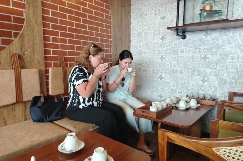 Two people sitting at a table with teapots and cups, in a room with a brick wall and patterned wallpaper. 5