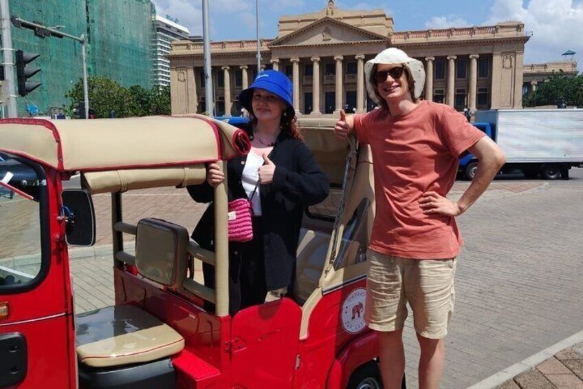 A red and beige tuk-tuk parked on a paved area with a building in the background. 2