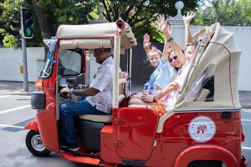 A red auto-rickshaw with passengers waving, a traffic light, and a white wall in the background. 1