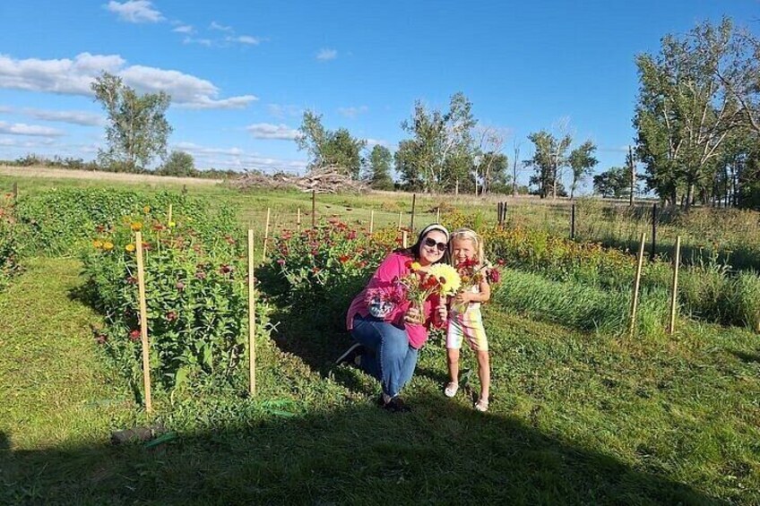 A person crouching in a field with a child holding flowers, surrounded by green vegetation and trees. 4