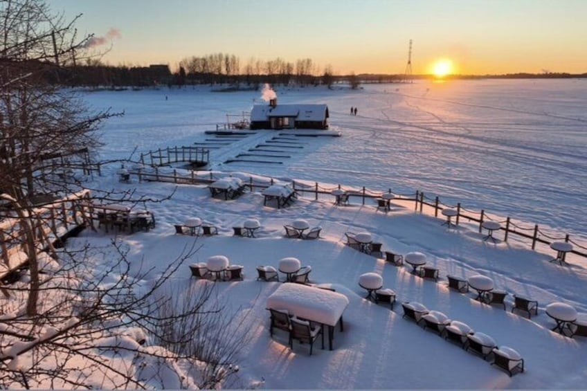 A snow-covered outdoor area with tables and chairs arranged for visitors, a building with a snow-covered roof, and a sunset in the background. 1