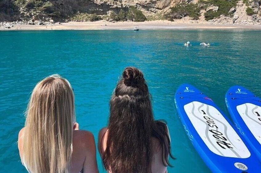 Two people facing a beach with clear blue water and a boat in the distance. 4