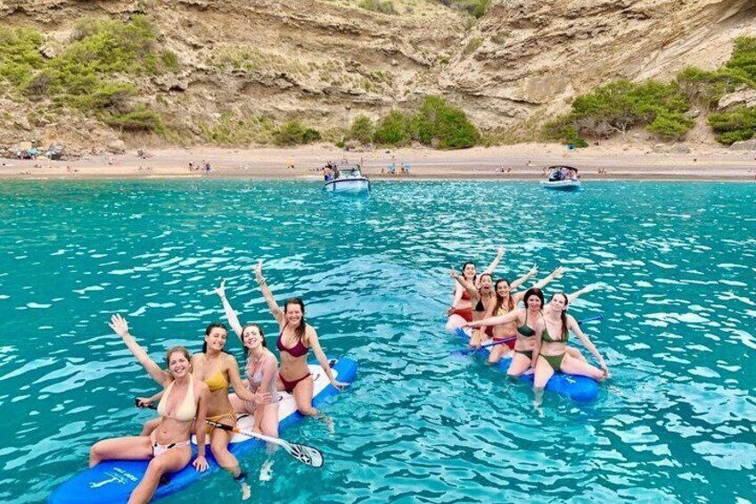 A group of people on inflatable rafts in a clear blue sea with a rocky coastline and boats in the background. 3