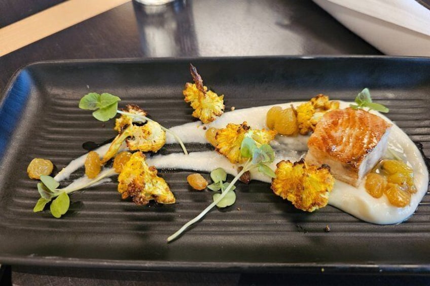 A plated dish with a piece of fish, cauliflower, and greens on a black plate. 1