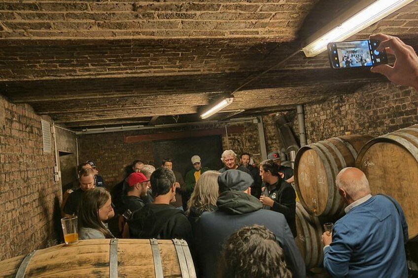 A group of people in a cellar with wooden barrels, engaged in conversation and tasting wine. 2
