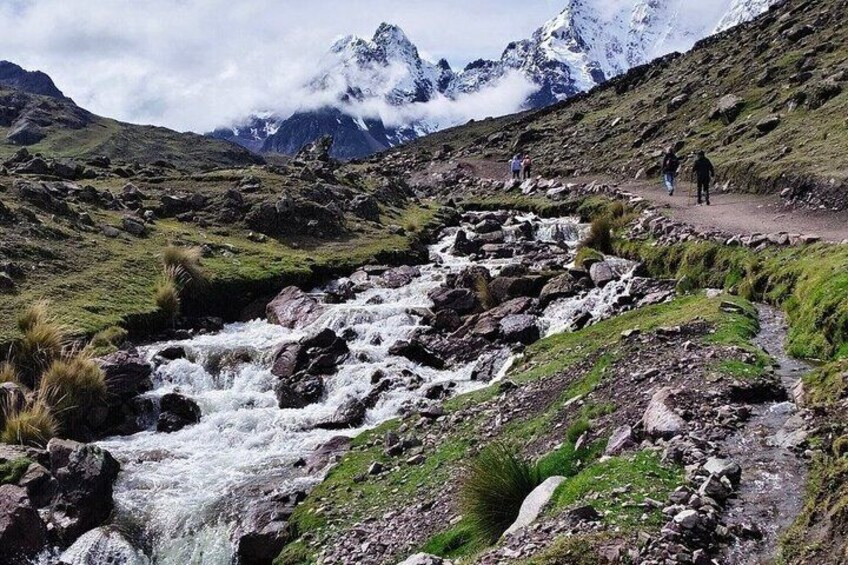 A mountainous landscape with a flowing stream, rocky terrain, and hikers on a trail. 4