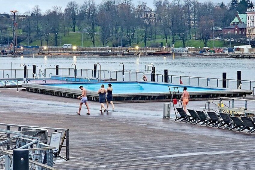 A wooden pier with a swimming pool, people walking, and a cityscape in the background. 3
