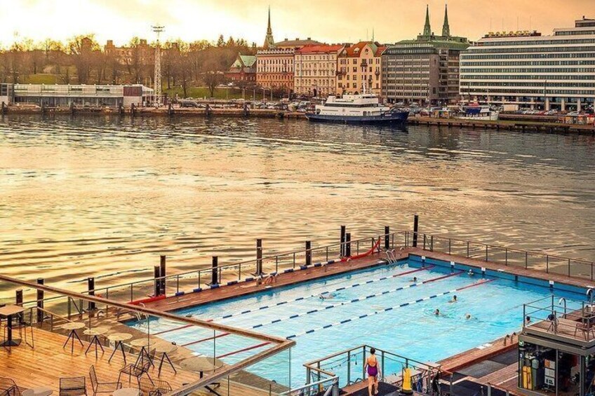 A swimming pool with a wooden deck, a cityscape with buildings and a church in the background, and a boat on the water. 1