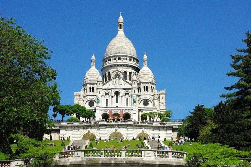 A large white domed building with multiple arches and a central dome, surrounded by greenery and a clear blue sky. 2