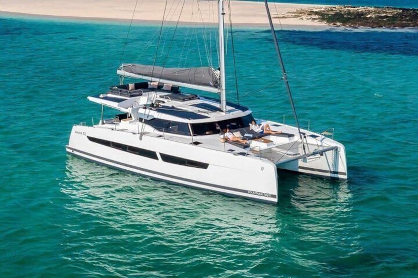 A white catamaran with a cabin and a sail, floating on turquoise water near a sandy beach. 1