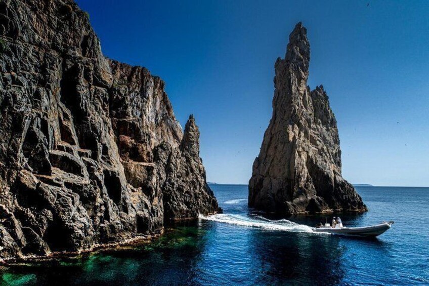 A boat on a clear blue sea with rocky cliffs and a tall rock formation. 1
