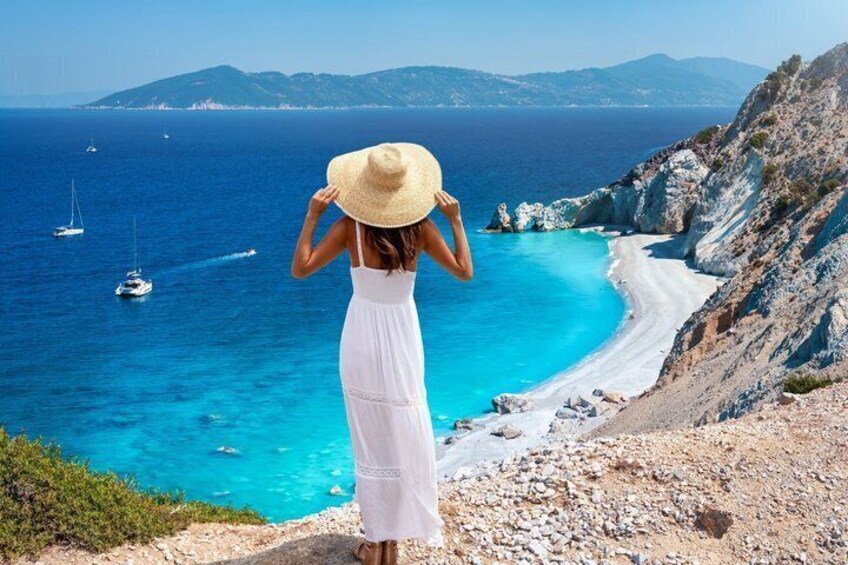 A woman in a white dress and straw hat overlooking a coastal landscape with clear blue waters, a sandy beach, and rocky cliffs. 5