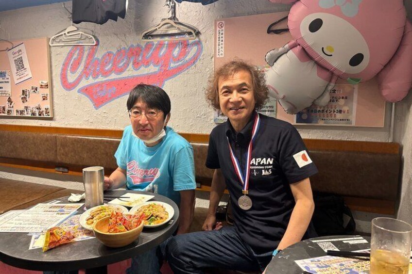 Two people sitting at a table with a bowl of food, a drink, and a menu. There is a wall with a 'Cheer Up' sign and a large Hello Kitty balloon. 2