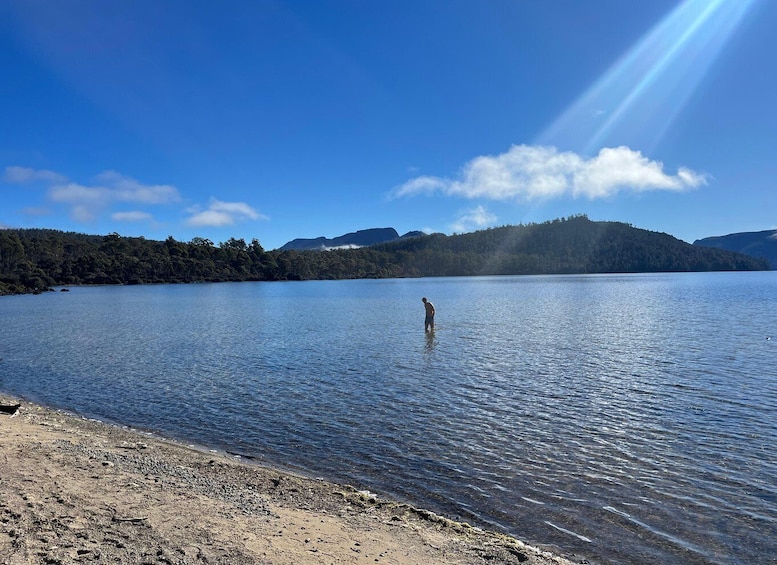 A calm lake with a person wading in the water, surrounded by forested hills and a clear sky. 5