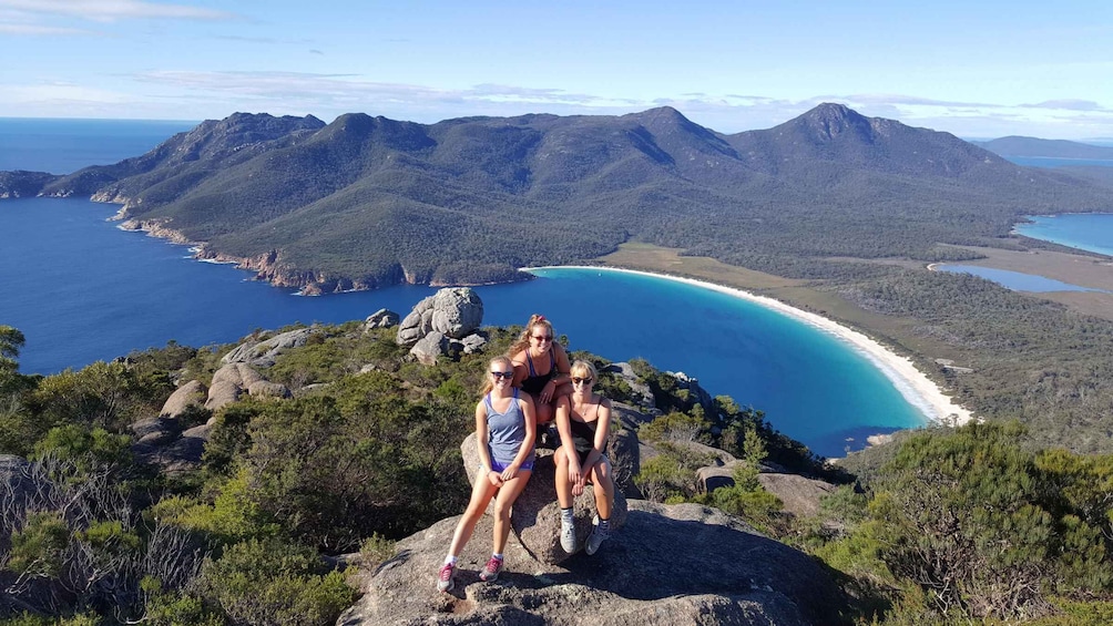 Three people sitting on a rock with a coastal landscape and mountains in the background. 3