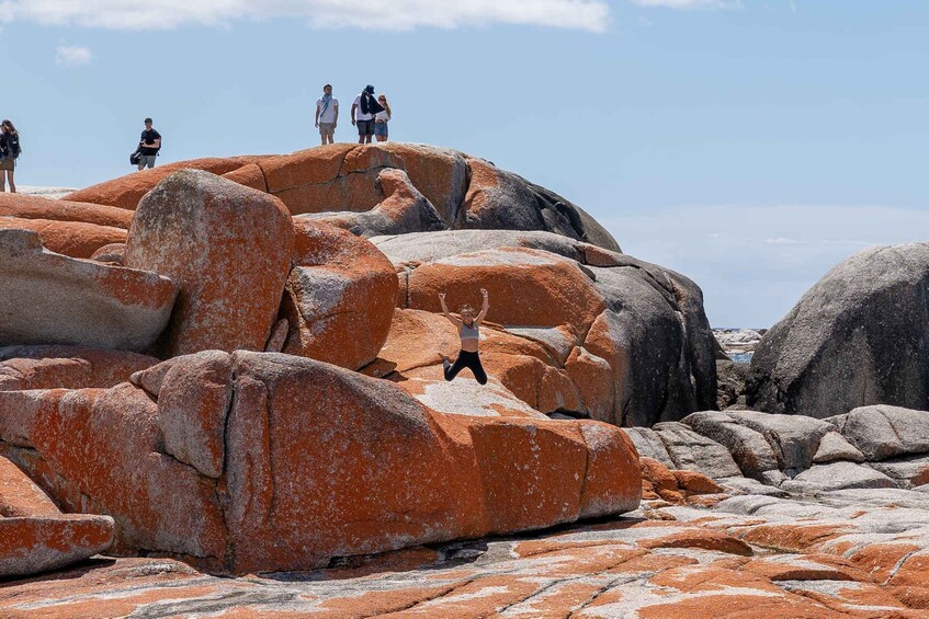 A person jumping on a large rock formation with others standing in the background. 1