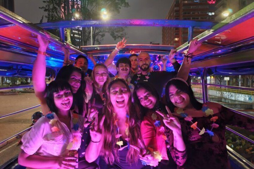A group of people on a brightly lit, multi-level bus with a cityscape in the background. 1