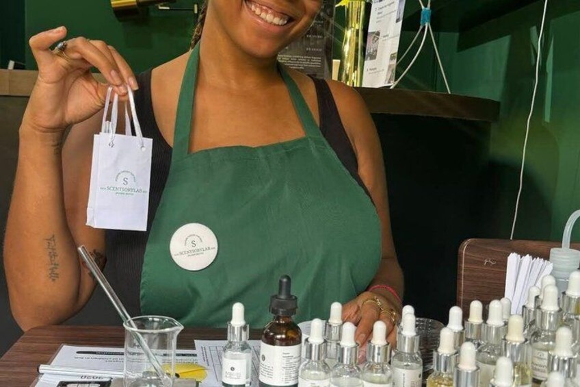 A person in a green apron holding a white bag, with various bottles and containers on a table in the foreground. 4