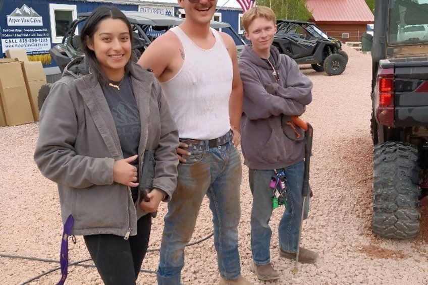 Three people standing in front of a vehicle, a building with a sign, and a red-roofed structure. 5