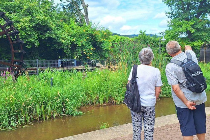 Two people standing by a canal with greenery and a wooden structure in the background. 3