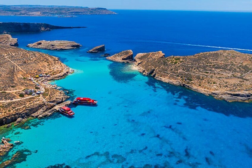 A coastal landscape with clear blue waters, rocky outcrops, and a few red boats anchored near the shore. 4