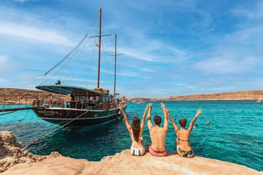 Three people sitting on a rocky ledge, facing a boat in the water. 1