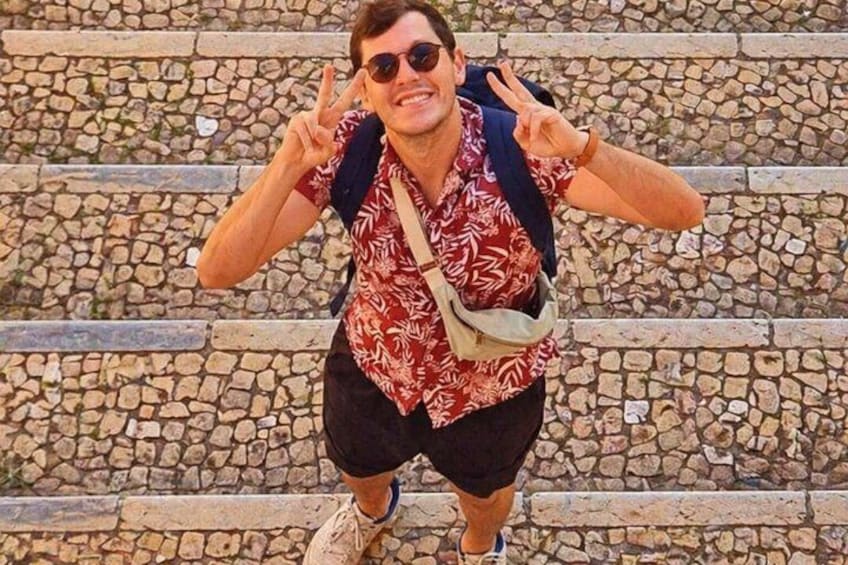 A person in a red patterned shirt, black shorts, and brown boots, making a peace sign with both hands, standing on a stone staircase. 5