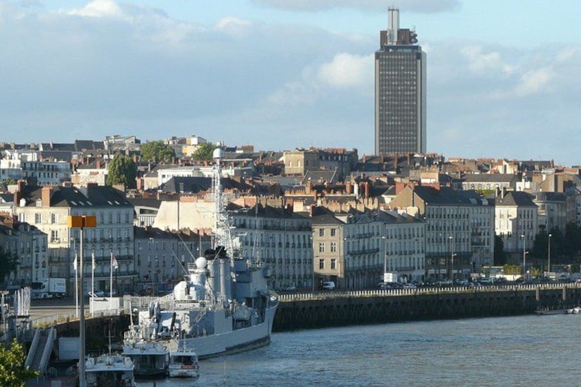 A large naval ship docked at a harbor, with a cityscape and a tall building in the background. 3