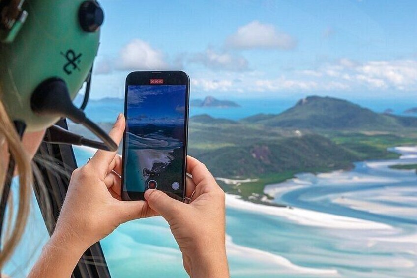A person holding a smartphone, taking a photo of a coastal landscape with mountains and a body of water. 4
