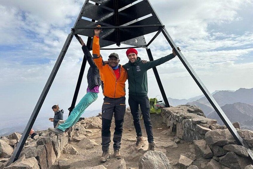 Two people standing on a rocky mountain peak with a metal structure in the background. 1
