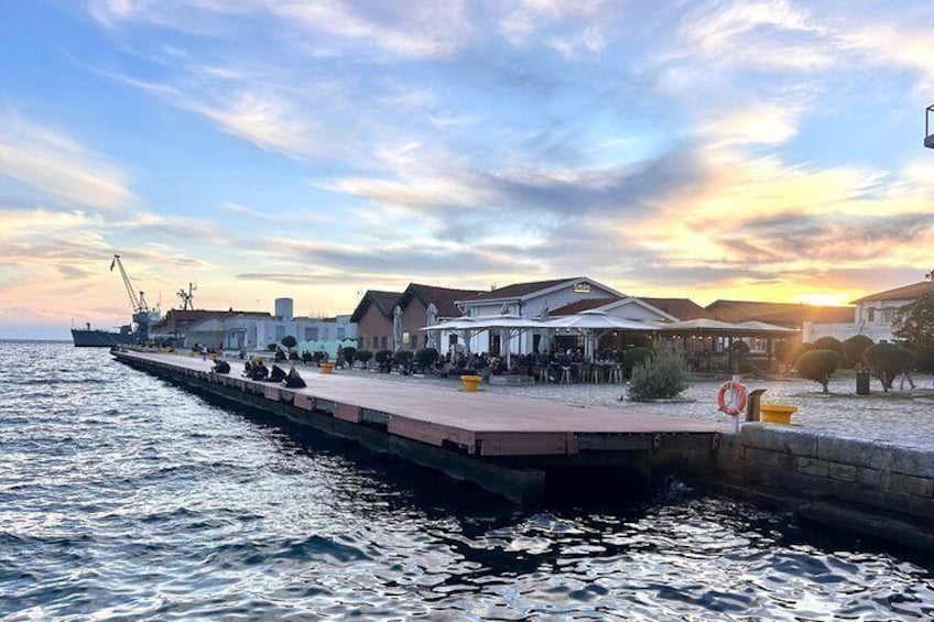 A waterfront promenade with buildings, a pier extending into the water, and a sunset in the background. 4