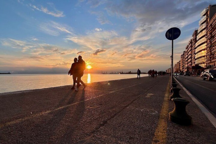A sunset over a waterfront promenade with people walking, a road with vehicles, and a building on the right. 2