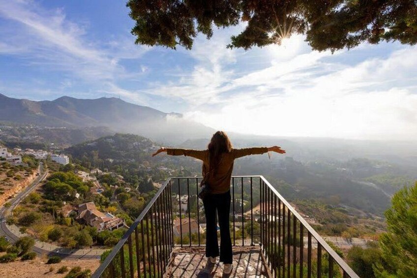 A person standing on a balcony with arms outstretched, overlooking a valley with buildings and mountains. 4
