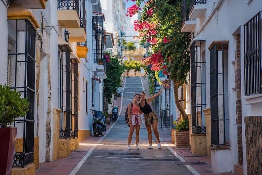 A narrow street with buildings on either side, a scooter parked on the side, and a tree with pink flowers in the middle. 3