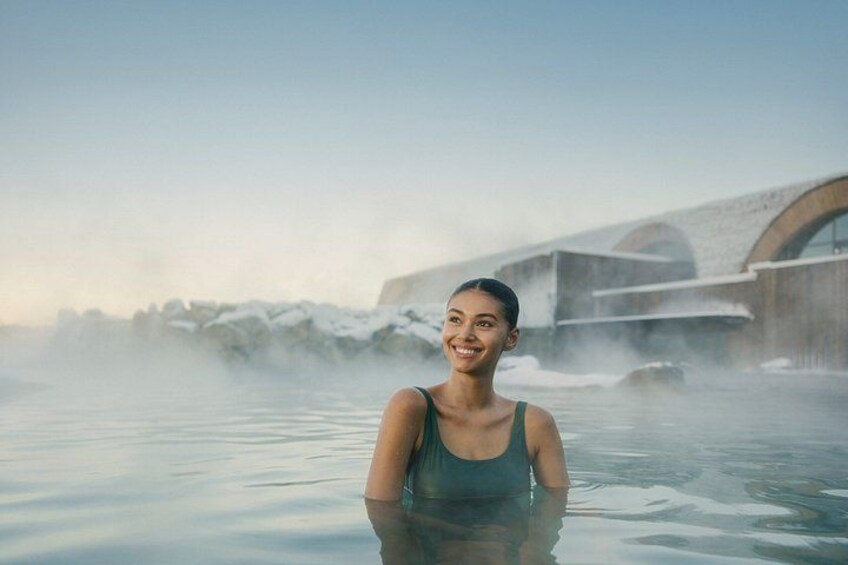 A person in a hot spring with steam rising, a bridge in the background, and a clear sky. 2
