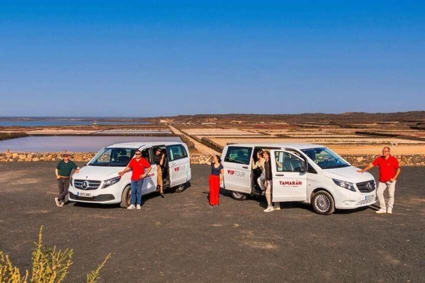 A group of people around a white van with 'TAMARAN' written on it, parked on a paved area with a clear sky and a barren landscape in the background. 1