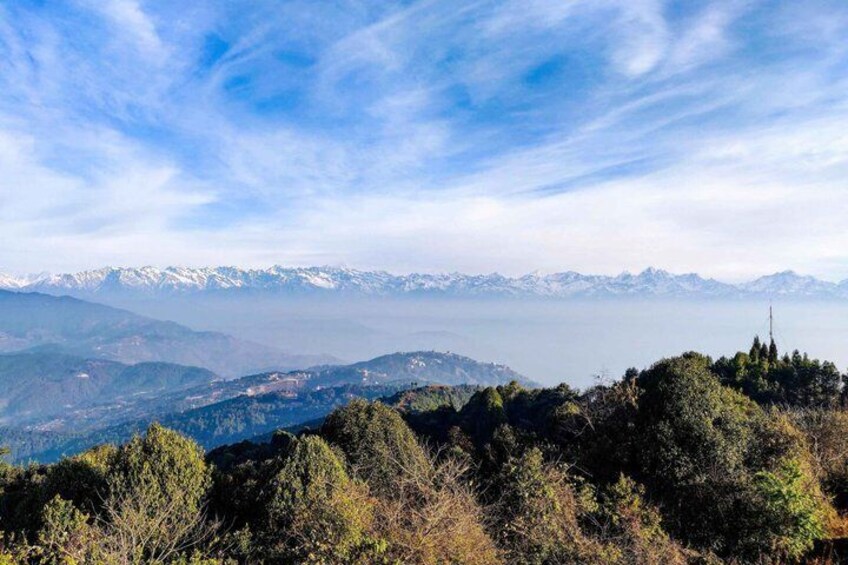 A mountainous landscape with snow-capped peaks in the distance, dense forest in the foreground, and a clear blue sky. 1