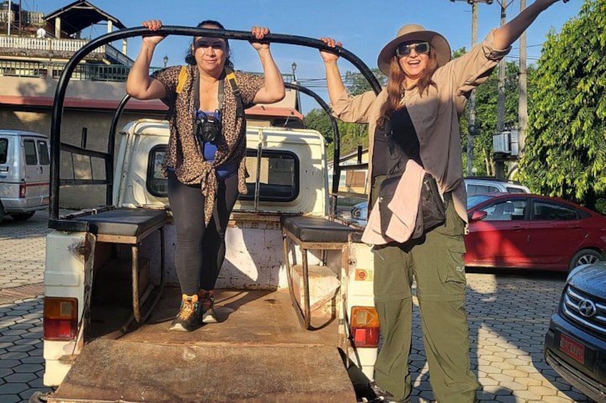 Two people standing on a vehicle's rear step, holding onto a metal bar. 3