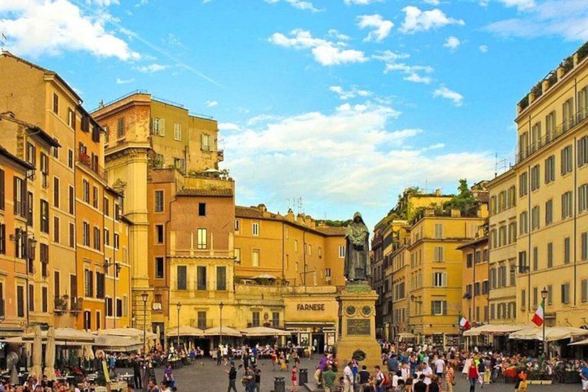 A crowded piazza with historic buildings, a statue, and a flag. 1