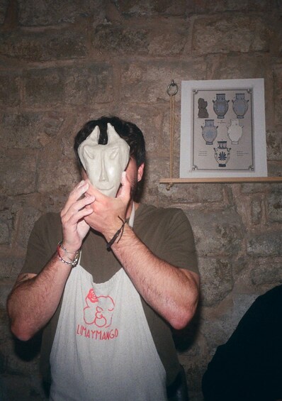 A person wearing a mask and an apron with 'LIMAYMANGO' text, standing in front of a stone wall with a framed picture of vases. 2